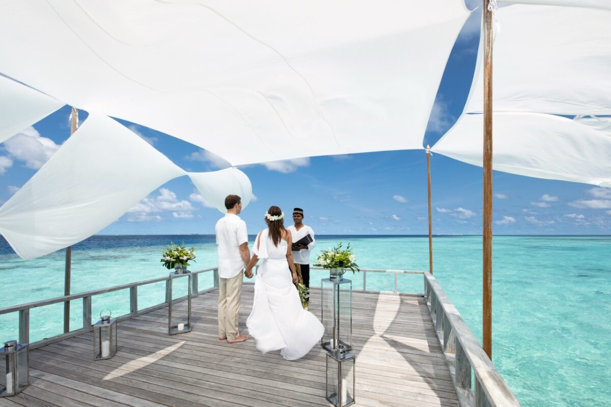 Mann und Frau in Hochzeitskleidung unter einem Stoffdach über dem Meer
