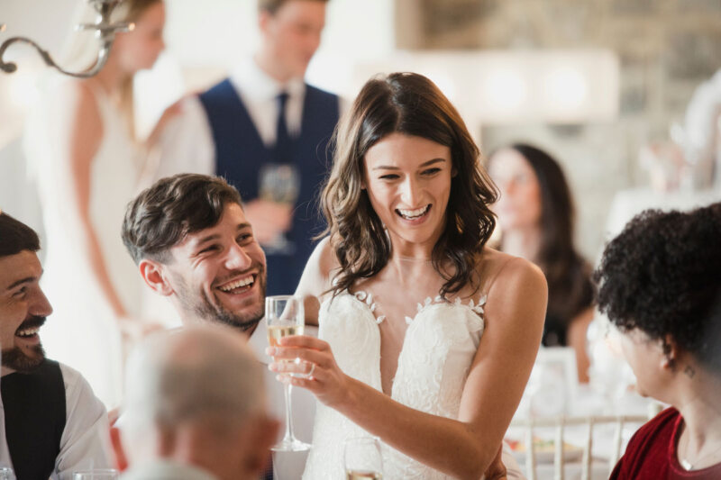Unterhaltung auf der Hochzeit: Eine fröhliche Braut und ein Bräutigam, die lachen, während sie auf ihre Gäste bei der Hochzeit anstoßen, umgeben von Familienmitgliedern in eleganter Kleidung. Das Paar trägt weiße Kleider mit Spitzendetails und hält Gläser Champagner oder Wein in der Hand.
