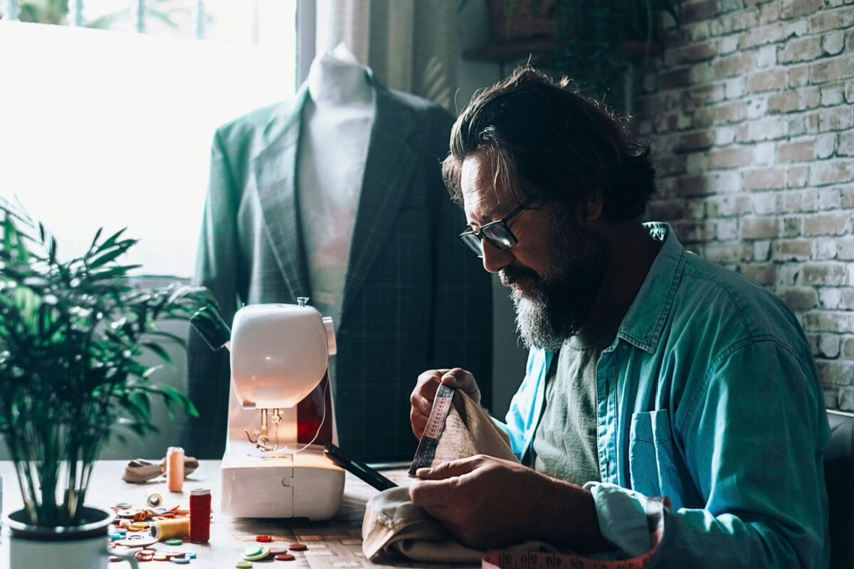 Hochzeitsanzug: Ein Mann mit Brille, Hemd und Bart sitzt an einem Tisch und näht an einer weißen Vintage-Mini-Designer-Nähmaschine von Singer neben einer Schaufensterpuppe in der Nähe eines Fensters. In der Nähe steht eine Pflanze in einer Vase und bunte Fäden sind im Raum verstreut. Warmes Licht von einer Lampe oben sorgt für eine gemütliche Inneneinrichtung.
