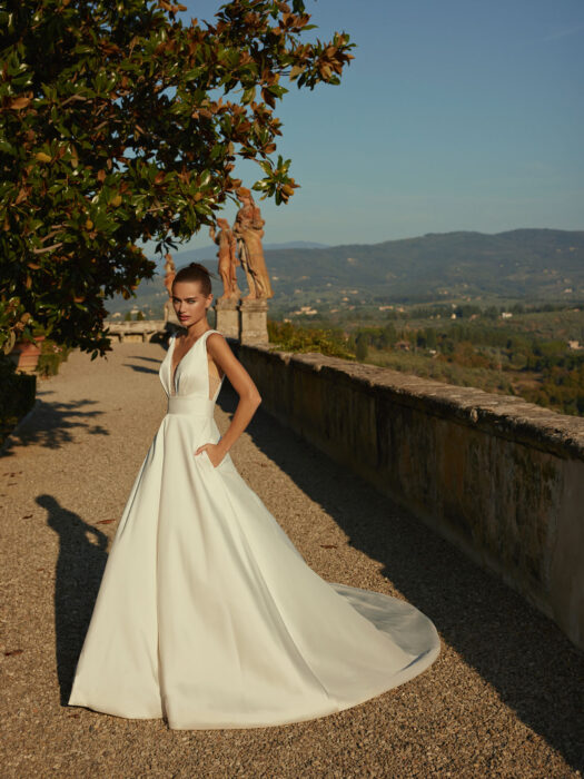 Hochzeitskleid aus glänzenden Mikadoseide im Prinzess-Schnitt, mit tiefen V-Ausschnitten vorn, hinten und an den Seiten sowie seitlichen Taschen und Schleppe von Modeca, Modell Deejay
